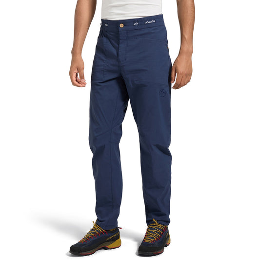 A person wearing La Sportiva Talus Pants Men's (2025) in navy blue and black hiking shoes with yellow and red accents stands against a white background, showing only the lower half of the body.