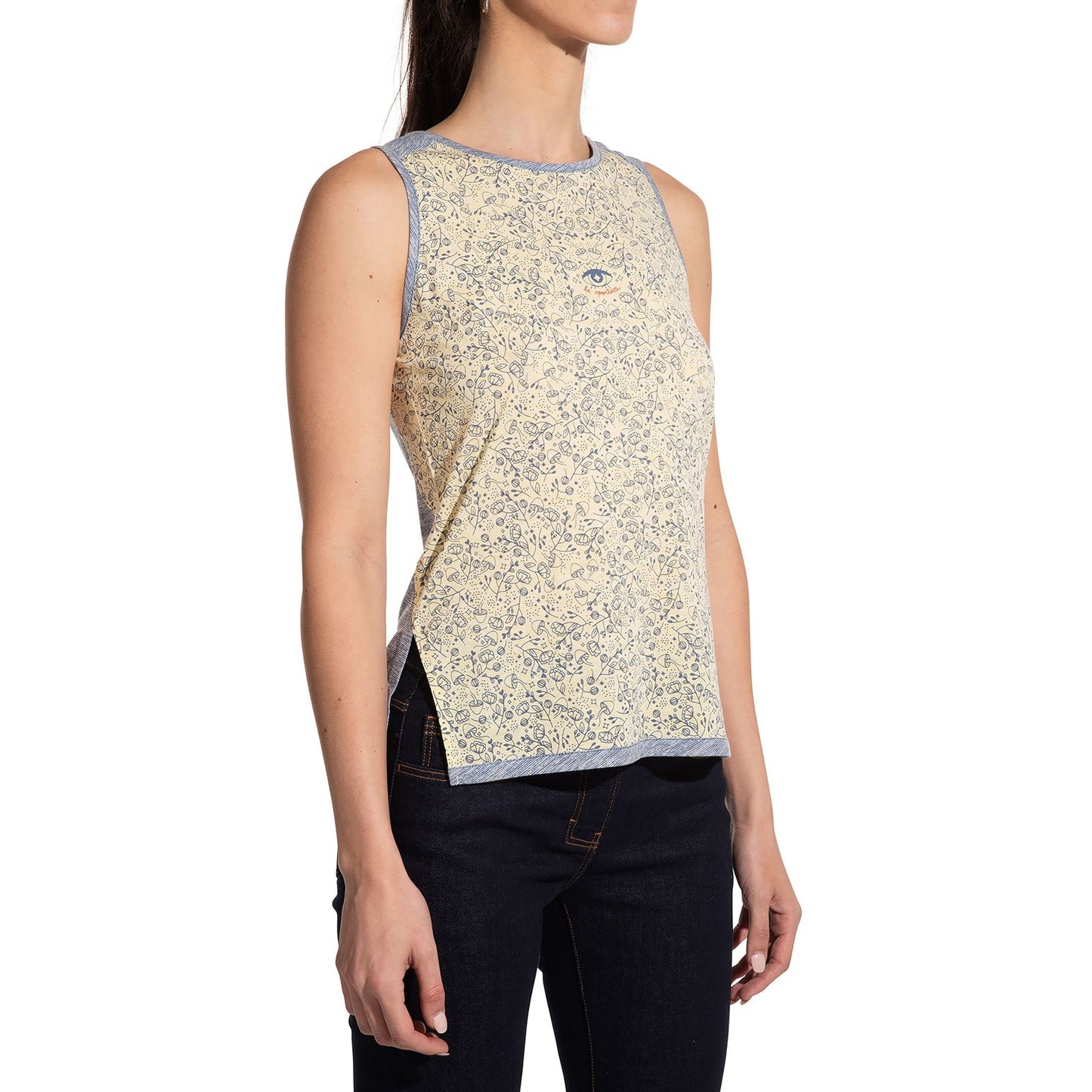 A woman stands against a white background wearing the La Sportiva Magic Tank Women's in sleeveless light yellow bamboo fabric with blue-gray trim, floral print, and a small eye graphic on the chest, paired with dark jeans.