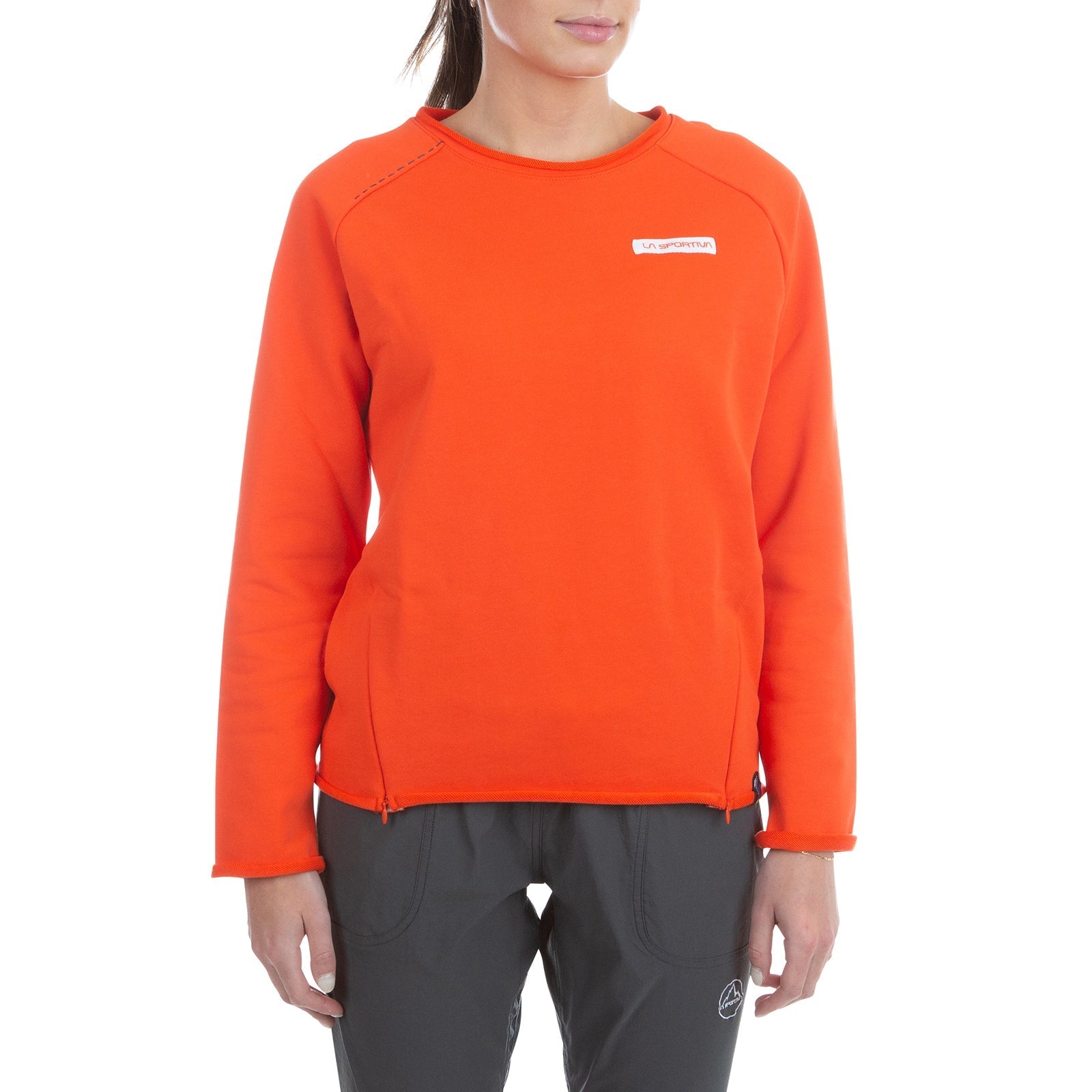 A woman wears the La Sportiva Tufa Sweater Women's (2025), an orange organic cotton climbing sweater with a small white chest logo and dark gray pants, standing against a white background.