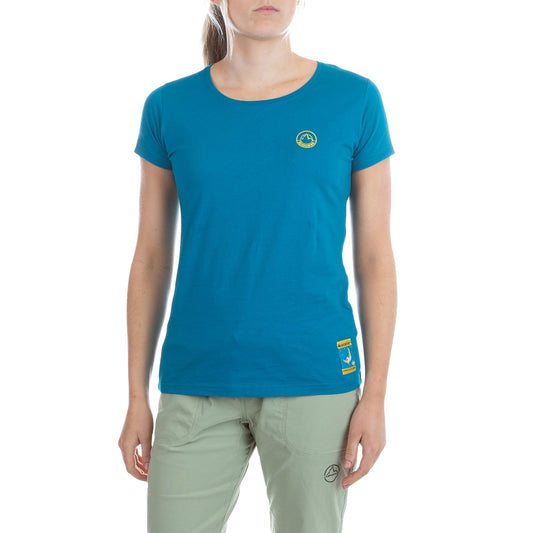 A woman wears the La Sportiva Climbing On The Moon T-Shirt Women's (2025) in teal with yellow logos, paired with light green pants, standing against a plain white background.