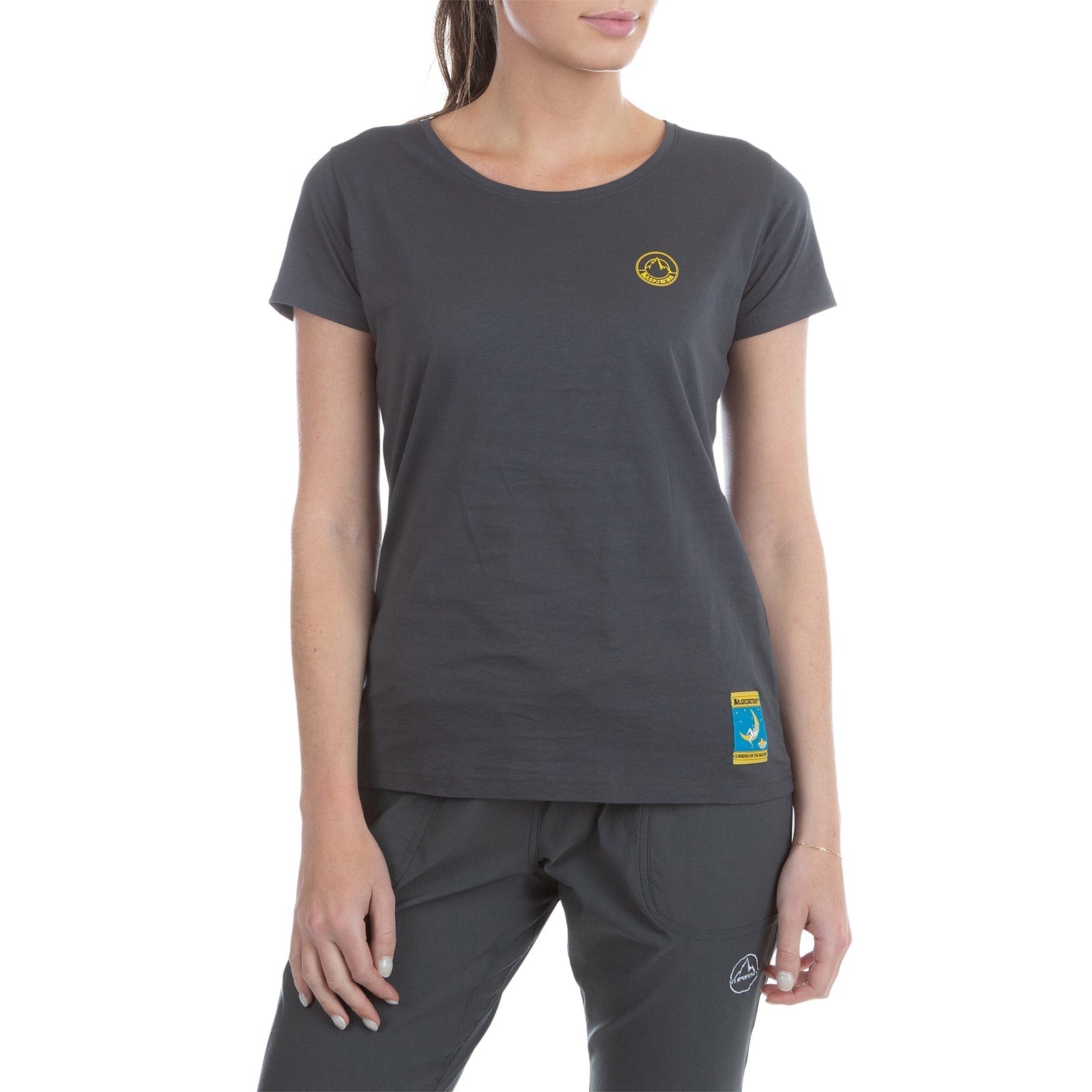 A woman wears the La Sportiva Climbing On The Moon T-Shirt Women's (2025) in dark gray with matching pants, featuring small yellow and blue logo patches. She stands relaxed against a plain white background, showcasing a subtle retro style.