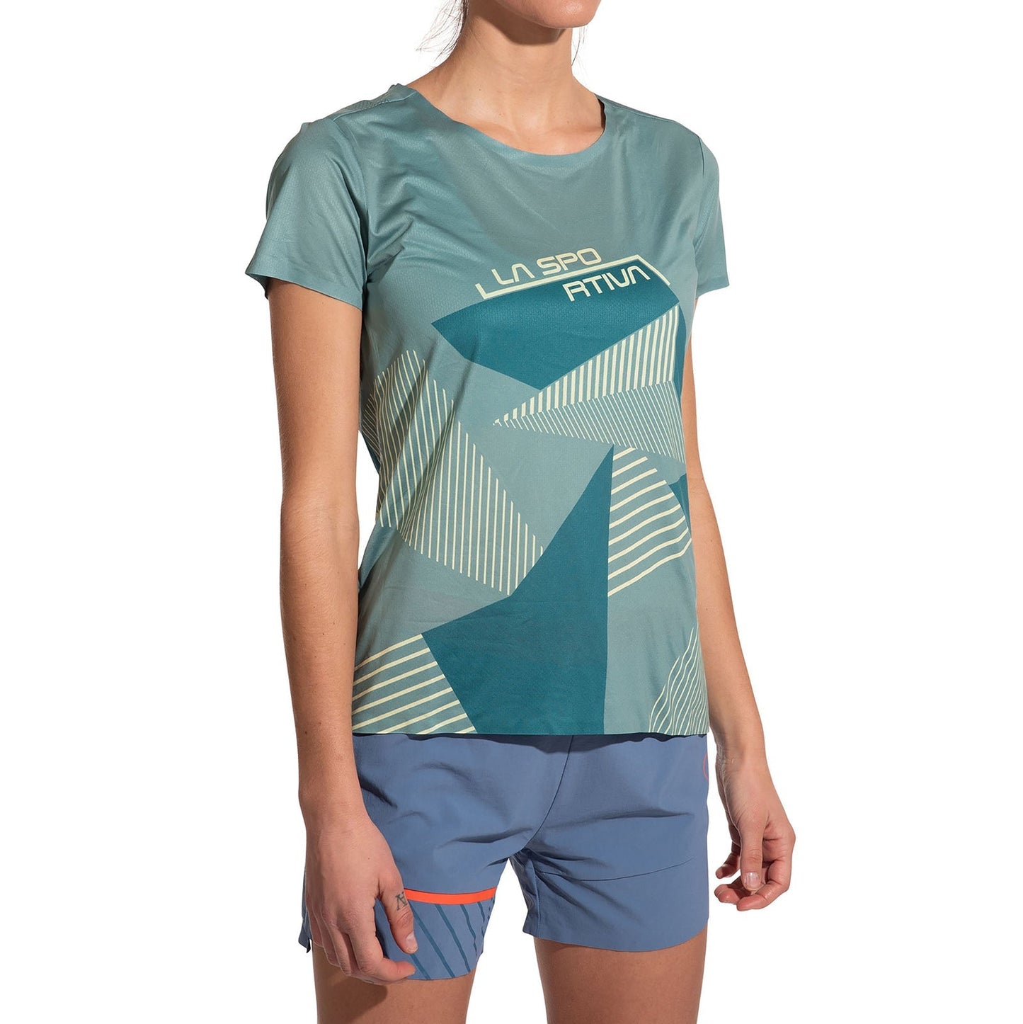 A woman wearing the La Sportiva Comp T-Shirt Women's (2025) in teal with a geometric pattern and blue athletic shorts stands against a white background, her head partially out of frame.
