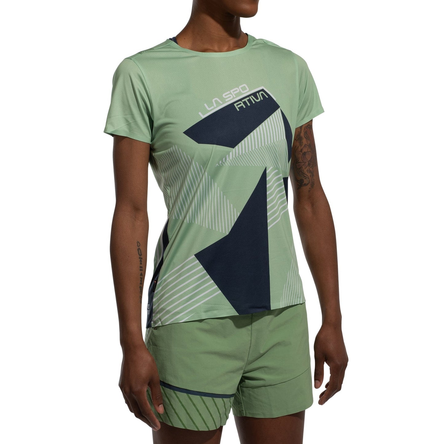 A woman wearing the La Sportiva Comp T-Shirt Women's (2025), a lightweight, light green athletic top with abstract dark blue and white geometric patterns, paired with matching green shorts, stands against a plain white background.