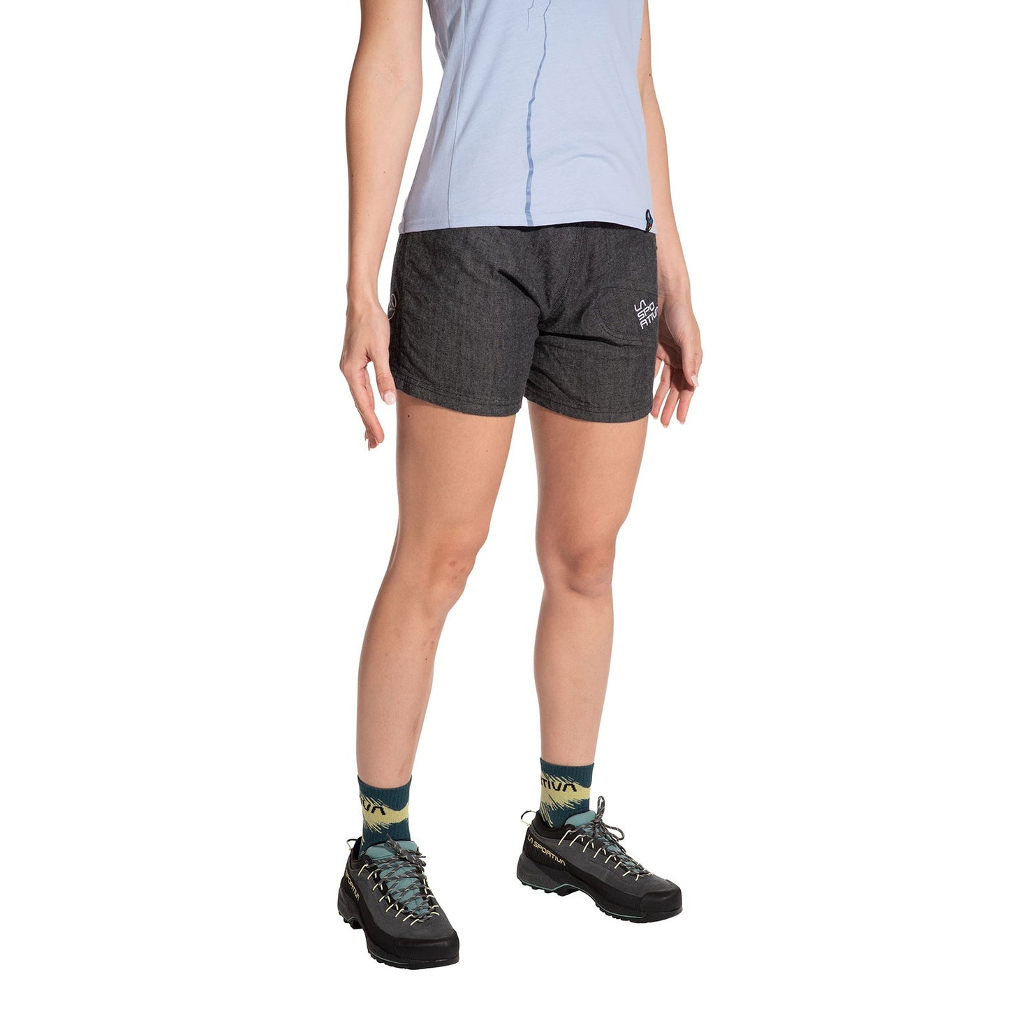 A person stands against a white background, shown from shoulders down, wearing the La Sportiva Sierra Rock Shorts Women's (2025), a light blue shirt, patterned socks, and black hiking shoes, highlighting comfort and functionality.