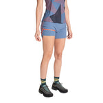 A person models La Sportiva Comp Shorts Women's (2025) in blue with a patterned athletic top, colorful socks, and black hiking shoes. The image captures them from shoulders to feet against a white background.