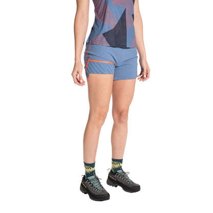 A person models La Sportiva Comp Shorts Women's (2025) in blue with a patterned athletic top, colorful socks, and black hiking shoes. The image captures them from shoulders to feet against a white background.