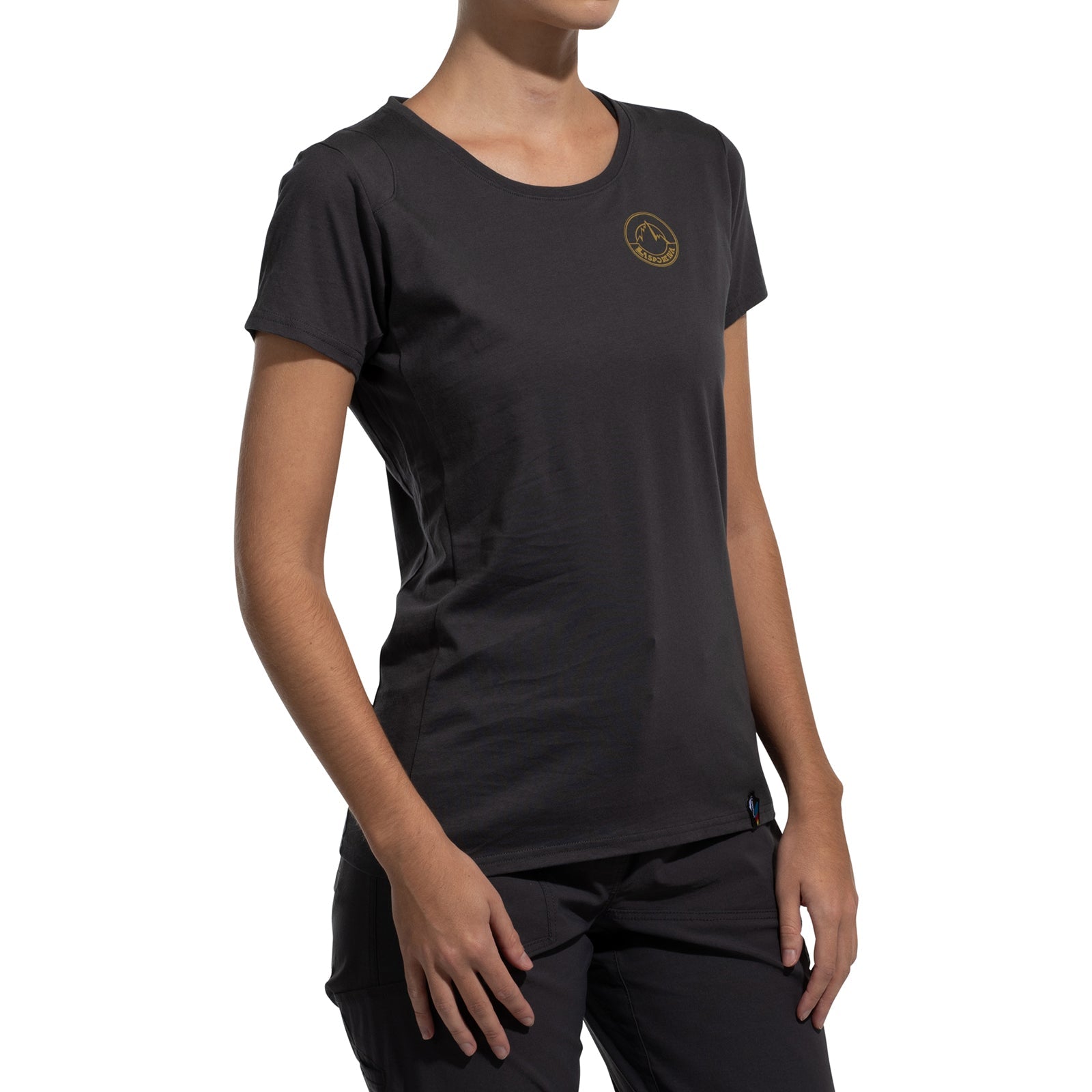A woman wears the La Sportiva Circle Logo T-Shirt Women's (2025) in black with black pants, standing relaxed against a plain white background. The fitted organic cotton tee is minimalist and features a small round logo on the upper left chest.