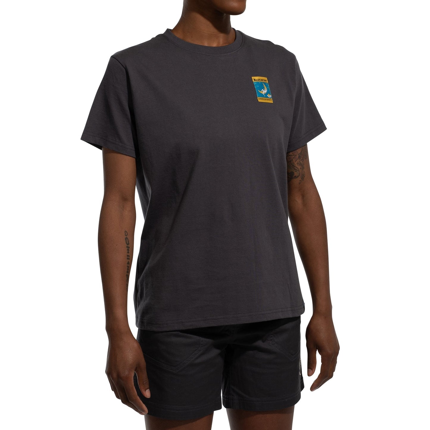 A woman wearing a La Sportiva Moon Climb T-Shirt Women's (2025), made of organic cotton with a small colorful chest patch, and black shorts stands against a white background.