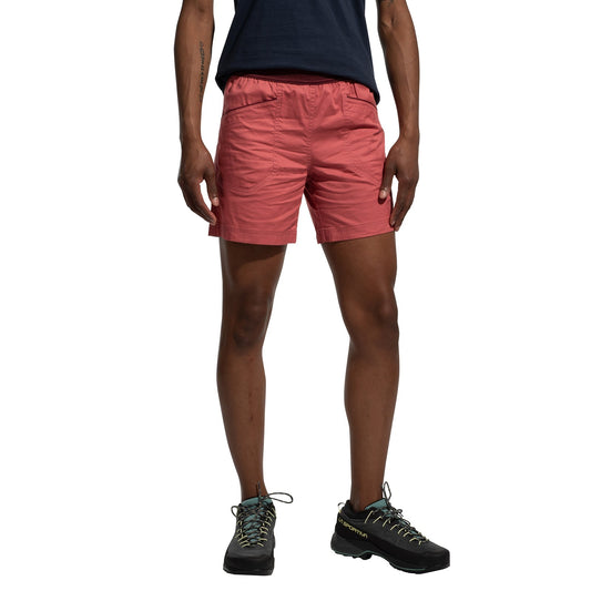 A person stands against a white background, shown from shoulders to feet, wearing a navy t-shirt, La Sportiva Roots Shorts Women's (2025) with a comfortable waistband in pink, and black hiking shoes; arms relaxed by their sides.