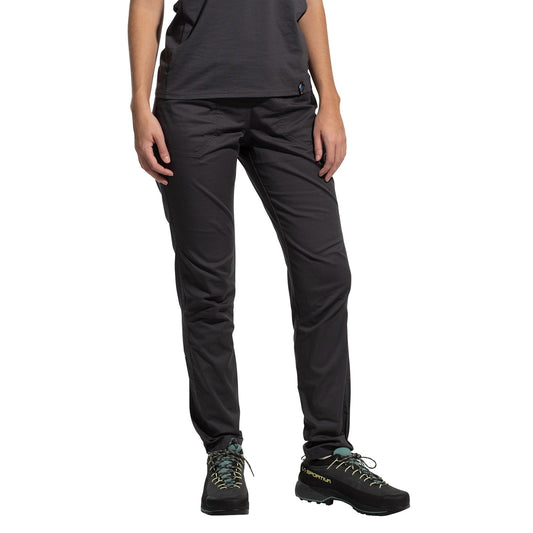 A woman wearing La Sportiva Roots Pants Women's (2025) in black stands against a white background. She also wears a dark gray organic cotton T-shirt and black hiking shoes with blue laces, shown from shoulders to feet.