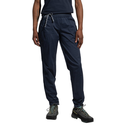 A person wearing La Sportiva Tundra Pants Women's in navy blue with articulated knees, black hiking shoes with green laces, and a matching navy shirt stands against a white background, left hand in pocket.