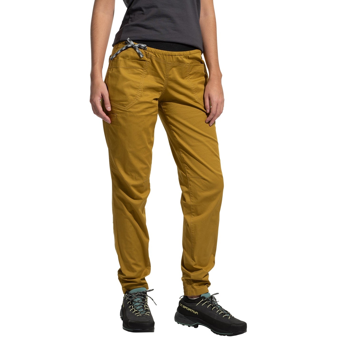 A person models the La Sportiva Tundra Pants Women's in mustard yellow with articulated knees, paired with a dark gray t-shirt and black hiking shoes. They stand against a white background, and their face is not shown.