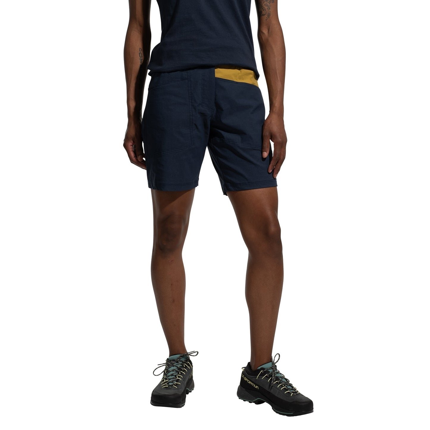 A person stands against a white background, wearing La Sportiva Mantra Shorts Women's (2025) in navy blue made from recycled fabric, paired with a matching t-shirt and black hiking shoes with light-colored laces.