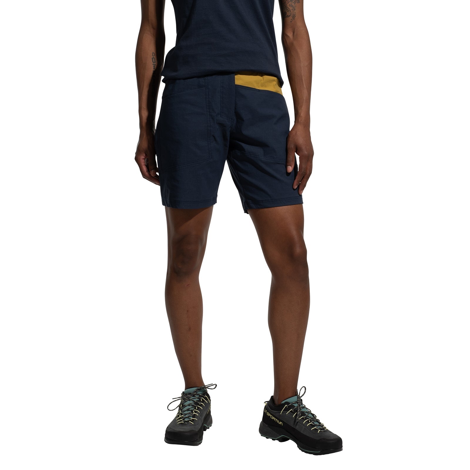 A person stands against a white background, wearing La Sportiva Mantra Shorts Women's (2025) in navy blue made from recycled fabric, paired with a matching t-shirt and black hiking shoes with light-colored laces.