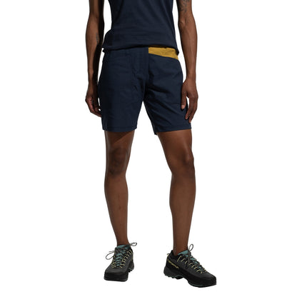 A person stands against a white background, wearing La Sportiva Mantra Shorts Women's (2025) in navy blue made from recycled fabric, paired with a matching t-shirt and black hiking shoes with light-colored laces.