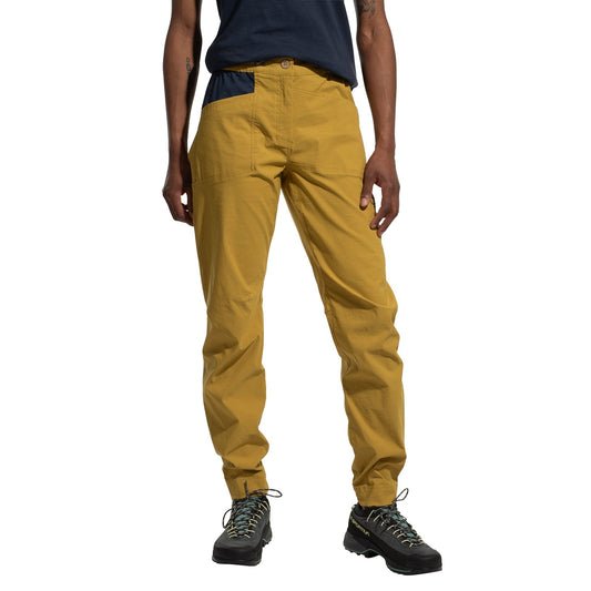 A woman in La Sportiva Mantra Pants Women's (2025) in mustard yellow with articulated knees and a navy blue pocket, paired with a dark t-shirt and black hiking boots, stands against a white background.