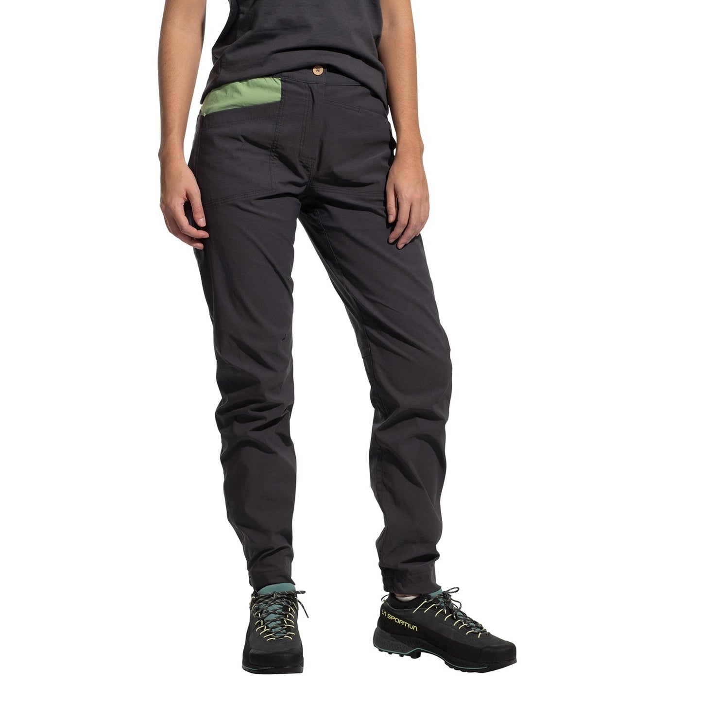 A person wears La Sportiva Mantra Pants Women's (2025) in dark gray with articulated knees and a green pocket, paired with black hiking shoes, standing against a white background. Photo features the lower torso and legs, arms relaxed at sides.