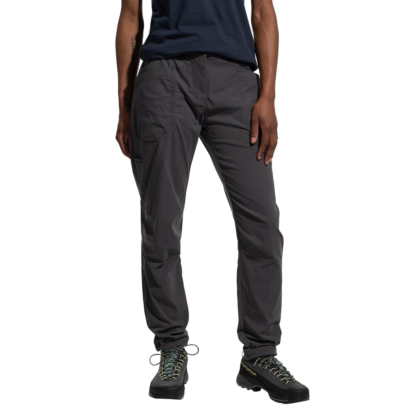 A person wearing La Sportiva Labyrinth Pants Women's (2025) with an adjustable waistband, a navy blue shirt, and black hiking shoes stands with one hand by their side and the other on their thigh. Photo is cropped at the shoulders.