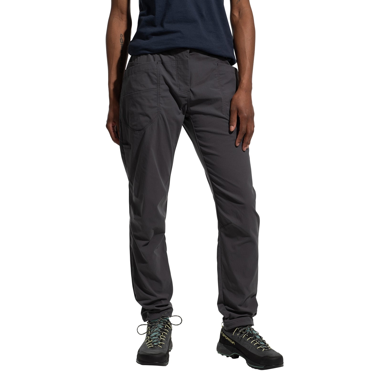 A person wearing La Sportiva Labyrinth Pants Women's (2025) with an adjustable waistband, a navy blue shirt, and black hiking shoes stands with one hand by their side and the other on their thigh. Photo is cropped at the shoulders.