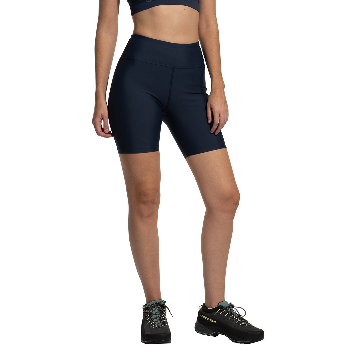 A person wears the La Sportiva Aerial Shorts Women's (2025) in navy blue with a gusseted crotch, paired with a matching sports bra and black hiking shoes, standing against a white background. Their face is not visible.