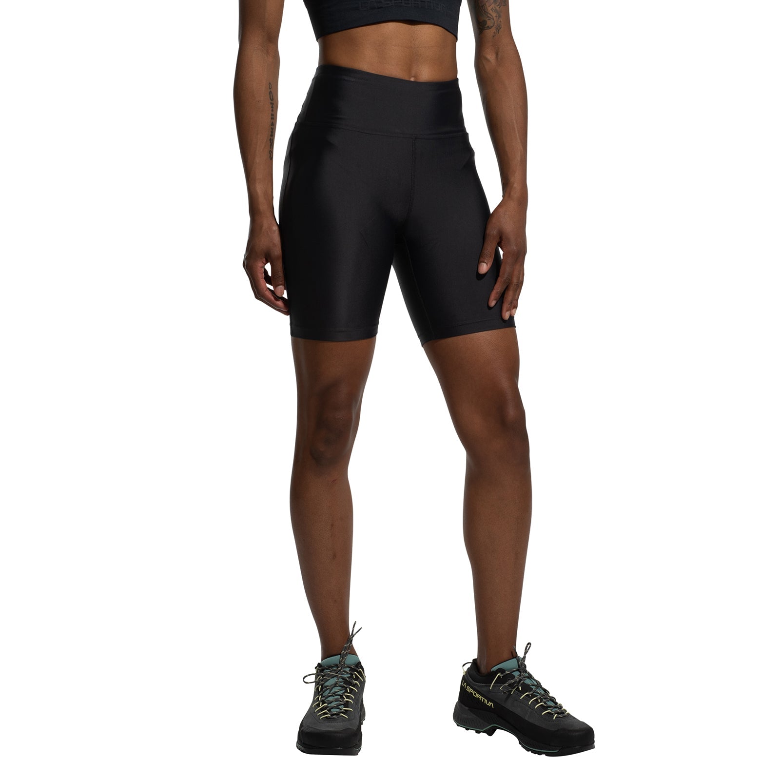 A person stands against a white background, arms relaxed, wearing a black sports bra, black La Sportiva Aerial Shorts Women's (2025) with a gusseted crotch and high waist, and black hiking shoes.