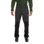 A person in La Sportiva Ikarus Pants Men's (2023), lightweight dark gray pants with zippered pockets, paired with black boots and a long-sleeve green shirt, stands against a white background.