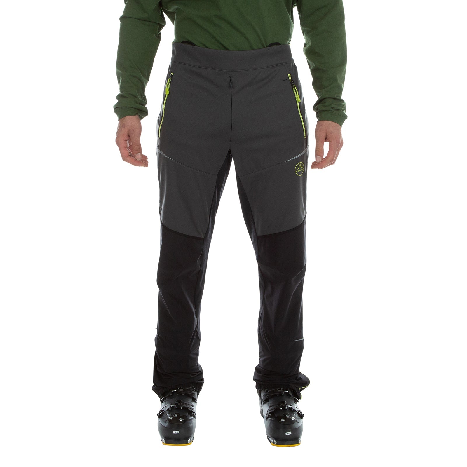 A person in La Sportiva Ikarus Pants Men's (2023), lightweight dark gray pants with zippered pockets, paired with black boots and a long-sleeve green shirt, stands against a white background.