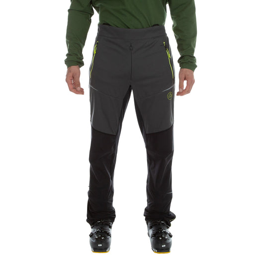 A person in La Sportiva Ikarus Pants Men's (2023), lightweight dark gray pants with zippered pockets, paired with black boots and a long-sleeve green shirt, stands against a white background.