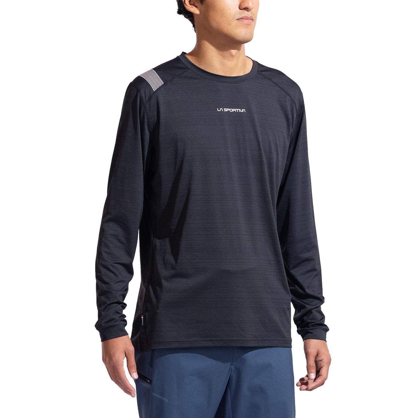 A person wearing the La Sportiva Tour Long Sleeve Men's shirt and blue pants stands against a white background, ready for active winter fun. This eco-friendly baselayer features reflective details for safety.