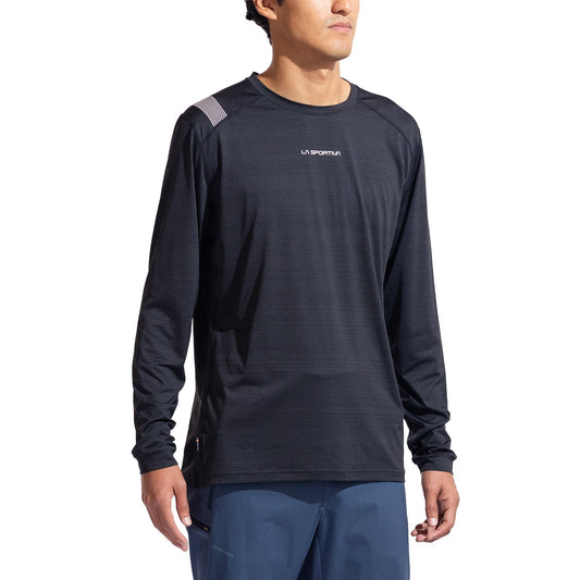 A person wearing the La Sportiva Tour Long Sleeve Men's shirt and blue pants stands against a white background, ready for active winter fun. This eco-friendly baselayer features reflective details for safety.