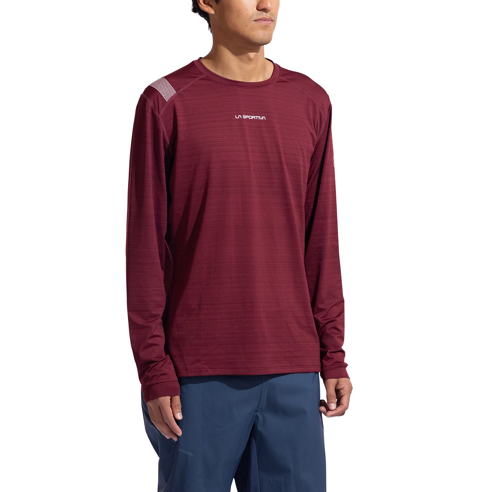 A man wearing the La Sportiva Tour Long Sleeve Men's shirt in maroon, styled as an eco-friendly baselayer with blue pants, stands against a white background, facing slightly to the side.