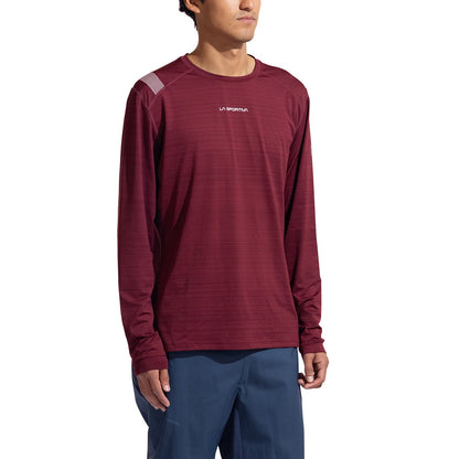A man wearing the La Sportiva Tour Long Sleeve Men's shirt in maroon, styled as an eco-friendly baselayer with blue pants, stands against a white background, facing slightly to the side.