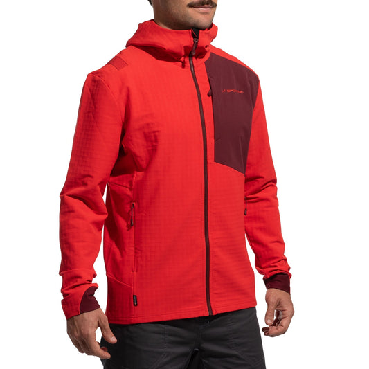 A man wearing the La Sportiva Descender Storm Jacket Men's (2025), a bright red softshell with maroon chest and cuff accents and a hood, paired with black pants, stands against a plain white background.