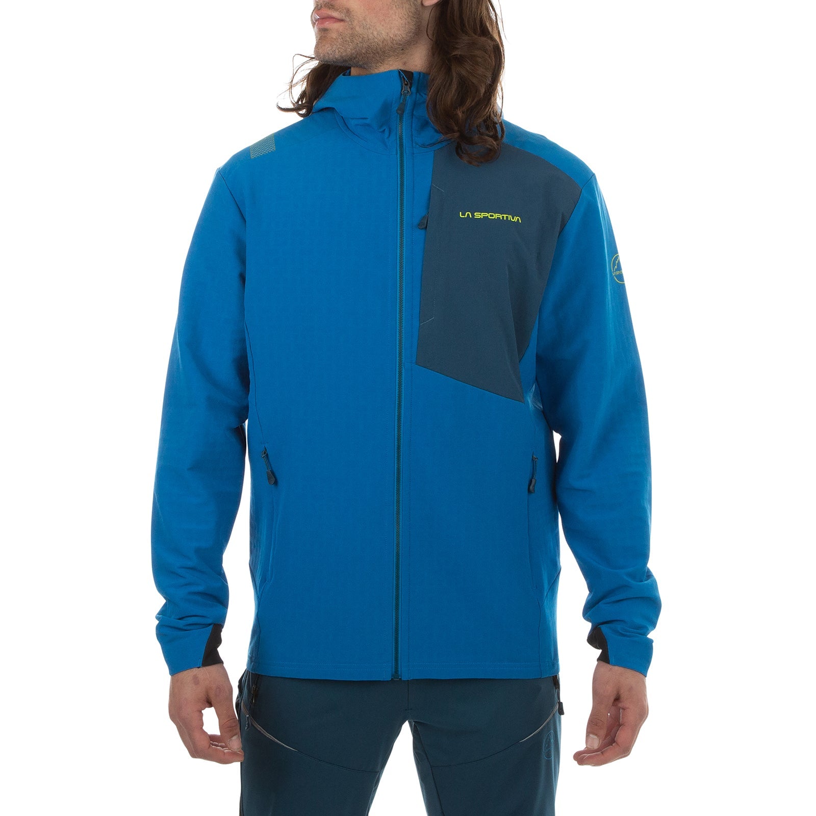 A person wears a bright blue La Sportiva Descender Storm Jacket Men's (2025) with a dark blue chest pocket, front zipper, hood, fleece interior, and zippered pockets. Hair partially obscures the face as they stand against a plain white background.