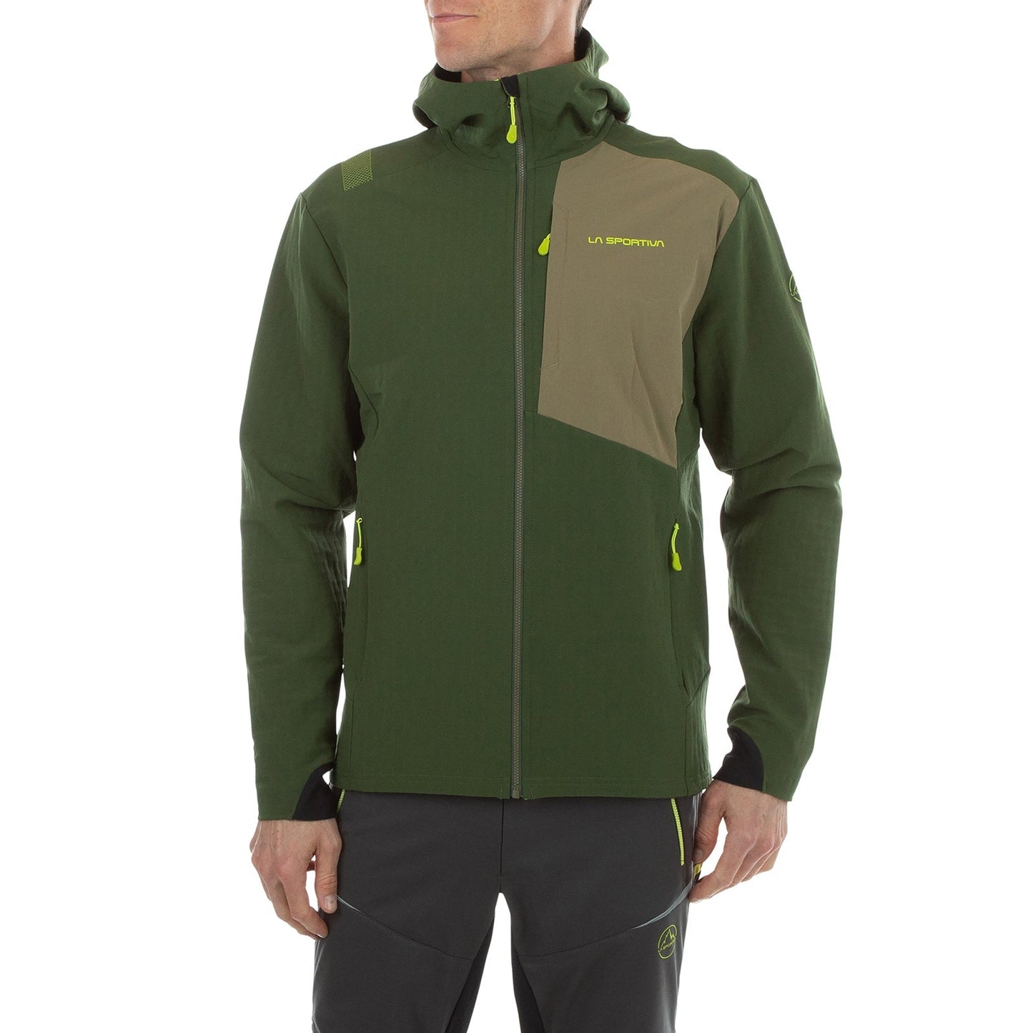 A man in the La Sportiva Descender Storm Jacket Men's (2025)—a dark green and olive softshell with fleece lining, yellow details, and chest logo—pairs it with black pants with yellow zippers, standing against a white background.