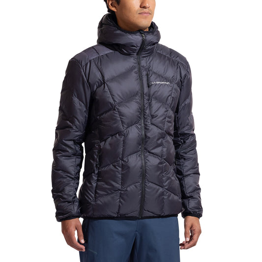A person wearing the La Sportiva Pinnacle Down Jacket Men's in dark gray with a quilted, hooded design and down insulation, paired with blue pants and standing against a plain white background.