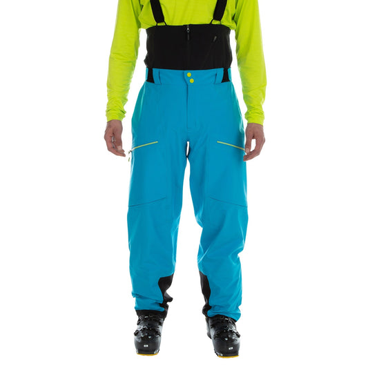 A person wears La Sportiva Chaser EVO Shell Bib Pants Men's in bright blue, paired with a neon yellow shirt and black ski boots, standing against a white background.