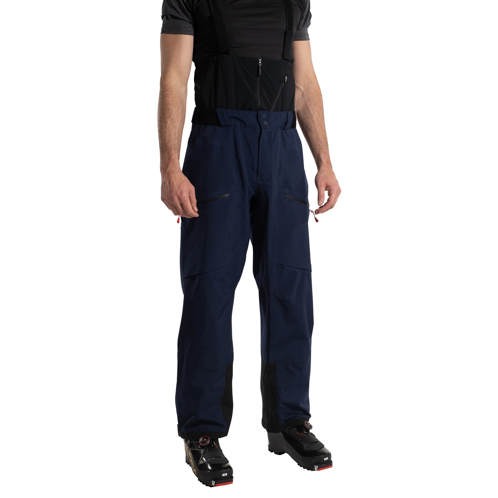 A person wears La Sportiva Chaser EVO Shell Bib Pants Men's in dark blue with black suspenders, paired with a black shirt and ski boots, standing against a white background. Their face is not visible.