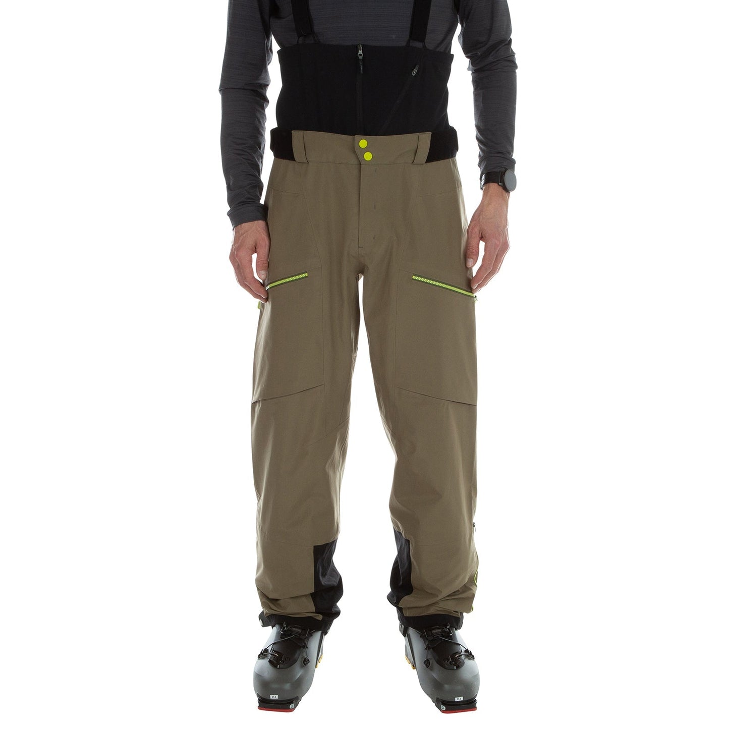 A person stands in black ski boots against a white background, wearing La Sportiva Chaser EVO Shell Bib Pants Men's in khaki-brown with a high waist, black panels, and yellow zippers.