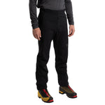 A person stands facing forward, showcasing the La Sportiva Ikarus Pants Men's in black with zippered pockets and a thigh logo, paired with a dark long-sleeve shirt and yellow-black hiking shoes. The image focuses on the lower body.