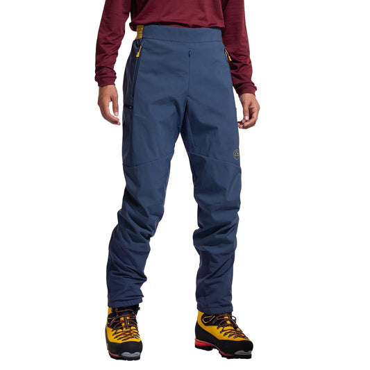 A person wears La Sportiva Ikarus Pants Men's—blue, breathable ski pants—paired with a maroon long-sleeve shirt and yellow hiking boots, standing against a white background. Photo is cropped at the shoulders.