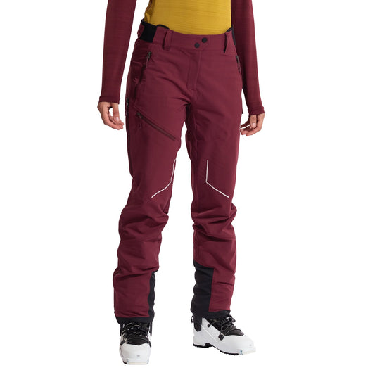 A person wears La Sportiva Excelsior Pants Women's—maroon breathable soft-shell pants with zippered pockets—plus black gaiters over white ski boots, a yellow top, and a red long-sleeve; ideal for ski-touring or mountaineering.