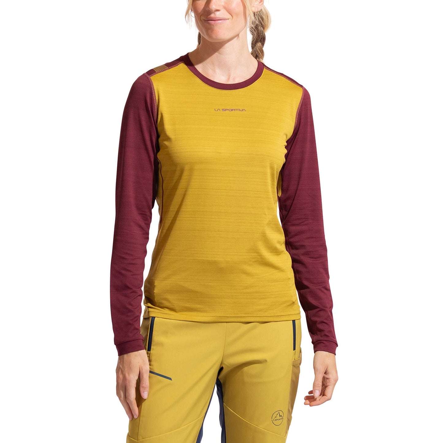 A woman stands against a white background wearing the La Sportiva Tour Long Sleeve Women's shirt in mustard yellow with burgundy sleeves, paired with matching mustard yellow women’s-fit pants.