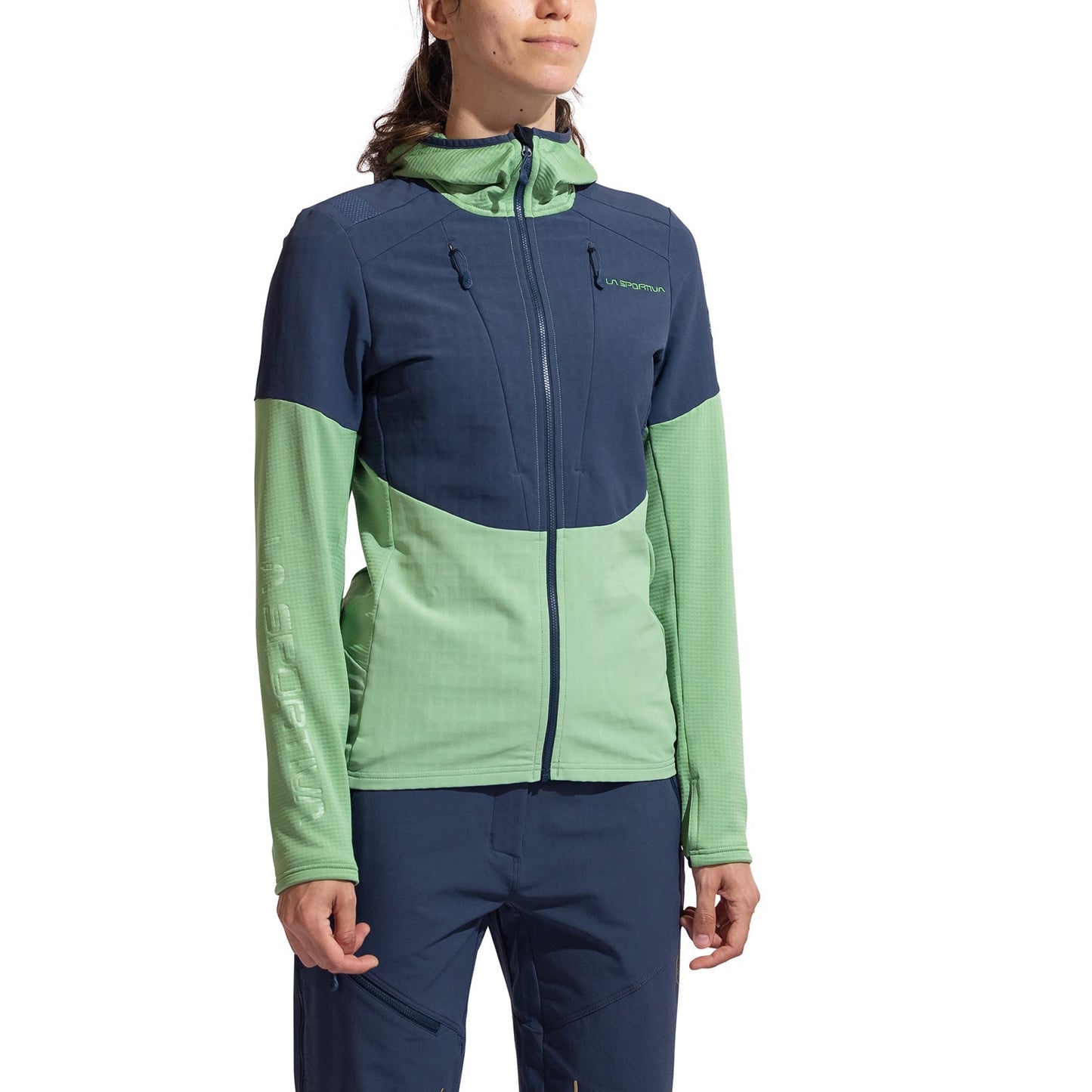A woman in a La Sportiva Session Tech Hoody Women's, featuring navy blue and light green panels with windproof body protection, pairs it with navy blue pants while standing against a white background.