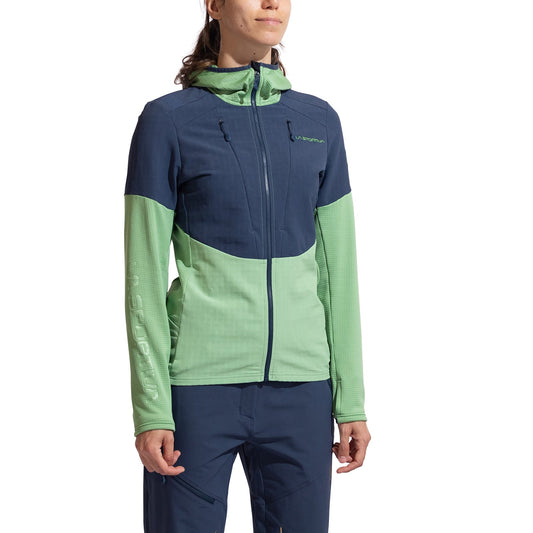 A woman in a La Sportiva Session Tech Hoody Women's, featuring navy blue and light green panels with windproof body protection, pairs it with navy blue pants while standing against a white background.