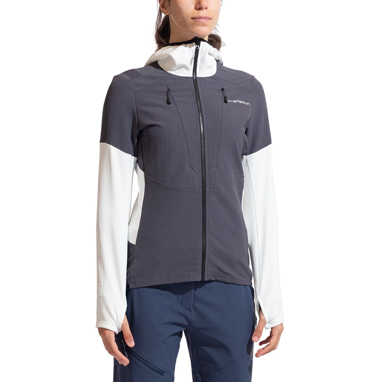 A woman wears the La Sportiva Session Tech Hoody Women's, featuring windproof protection, a color-blocked design (dark gray front/sleeves, white side panels/hood), two chest pockets, a front zipper, and navy blue pants.