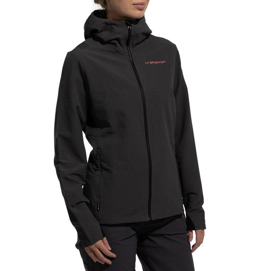 A person wearing the La Sportiva Descender Storm Jacket Women's (2025), a black softshell with fleece interior, side pockets, and front zipper, stands against a white background. The La Sportiva logo is on the chest; their face is partly out of frame.
