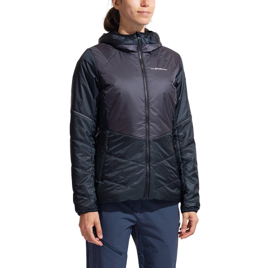 A person wearing a dark, hooded La Sportiva Mythic Primaloft Jacket Women's with long sleeves and navy blue pants stands against a white background. The insulated jacket has a subtle sheen and displays the La Sportiva logo on the left chest.
