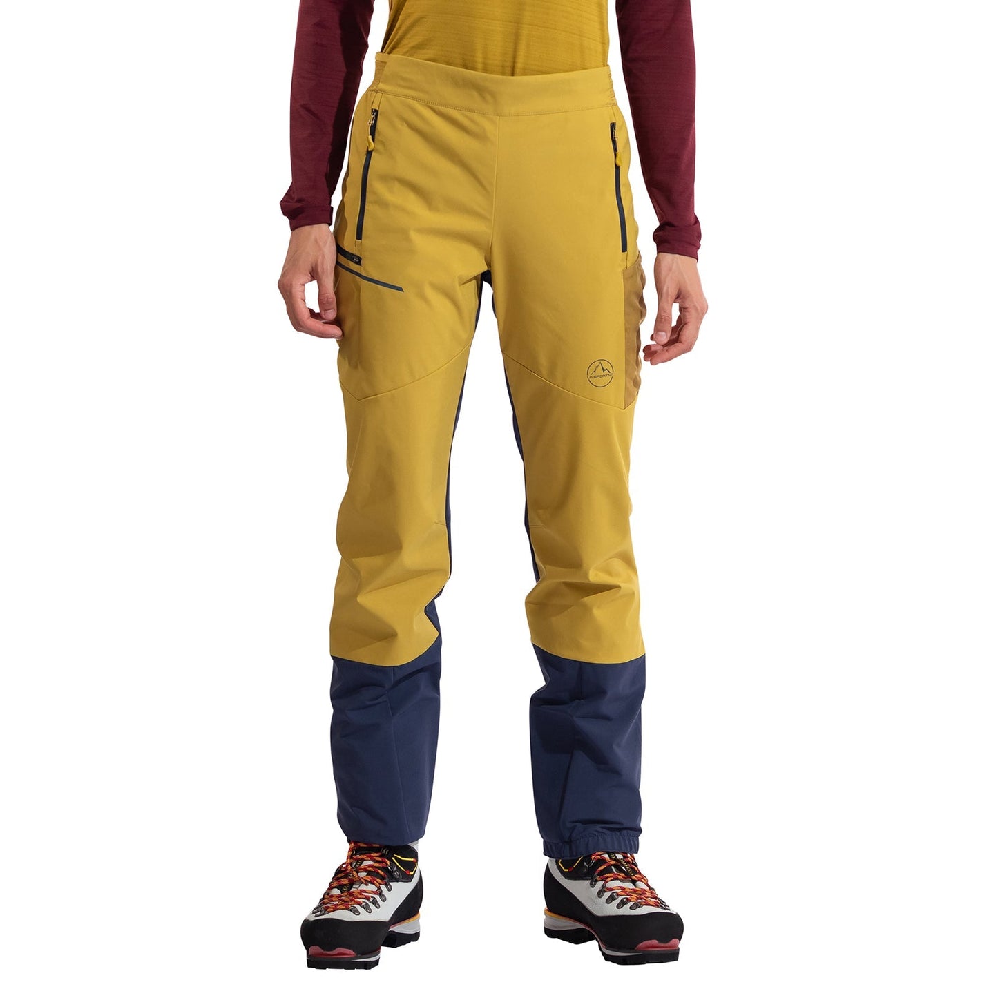 A person is wearing La Sportiva Ikarus Pants Women's—yellow, lightweight, breathable with navy blue lower legs—paired with a yellow shirt, maroon long-sleeve top, and hiking boots, standing against a white background.