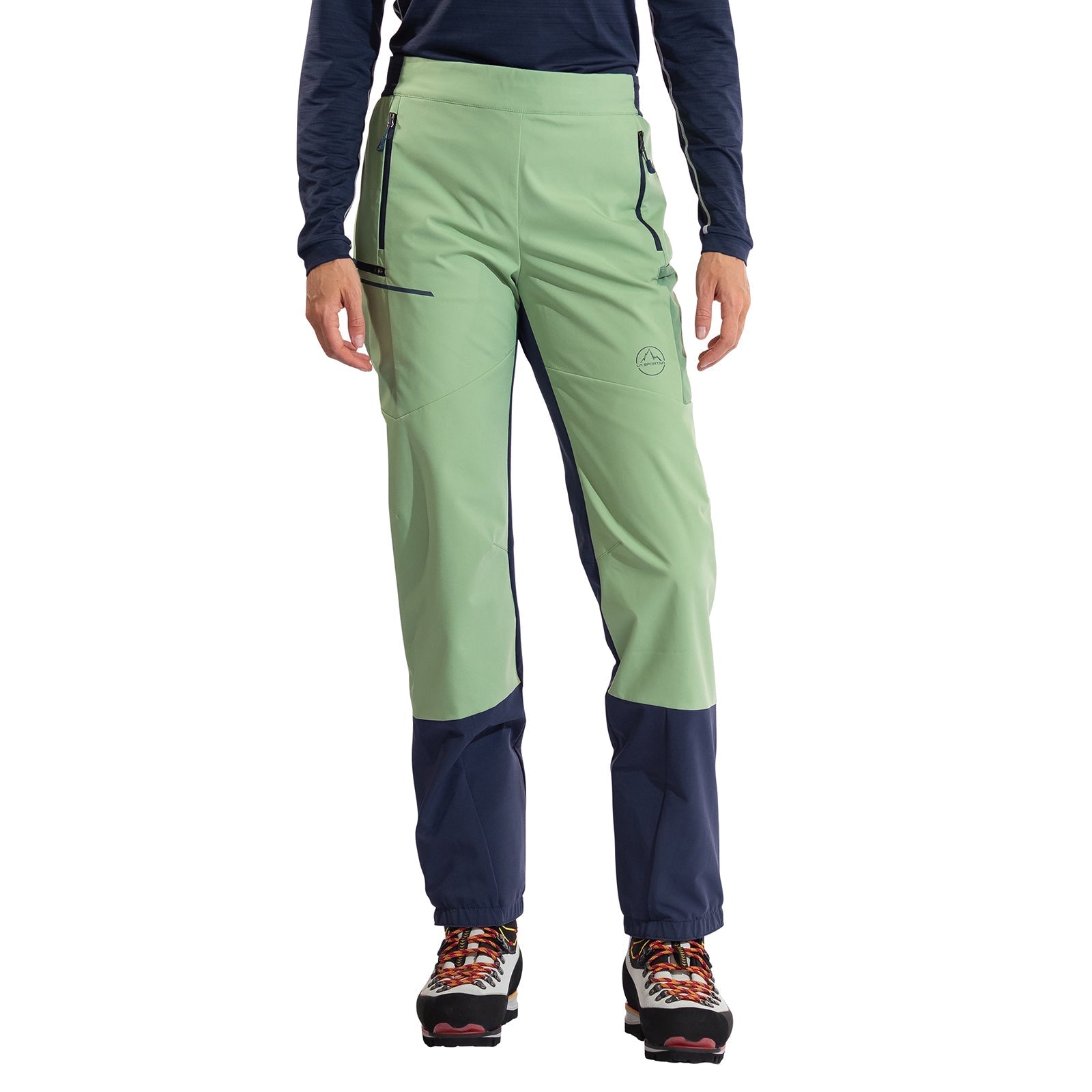 A person wearing La Sportiva Ikarus Pants Women's in light green and dark blue with zippered pockets, a navy long-sleeve top, and multicolored hiking shoes stands against a white background.